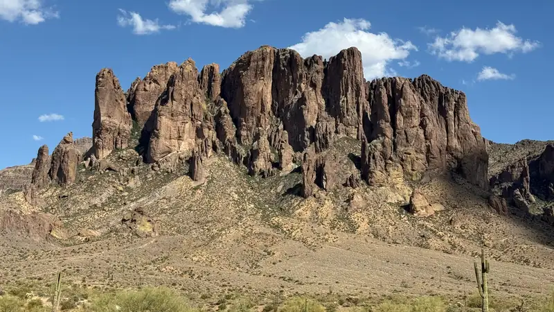 The cliffs of the Superstition Mountains above a desert slope dotted with saguaros, under a blue sky with a few clouds