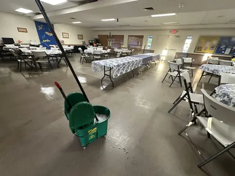 A hall set with round and long tables under flowered cloths and white folding chairs, its sealed floor wet and shining around a green mop bucket
