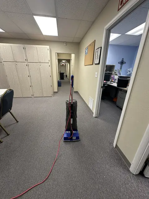 An upright vacuum with a red cord standing on grey carpet in an office corridor, beside the open door of an office with a desk