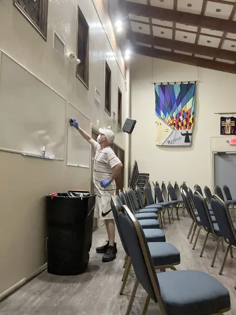 A crew member in a cap, a face mask and blue gloves reaches up to wipe a whiteboard in a hall with rows of stacking chairs and a quilted banner on the wall